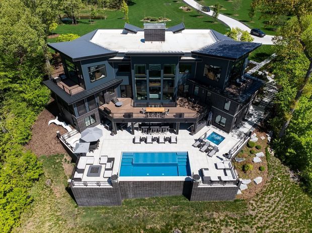 Aerial view of a large, dark-clad multi-storey residence set within a landscaped suburban or rural property. The home has multiple angular roof forms, extensive outdoor terraces and balconies, a central glazed vertical section, and a large rectangular swimming pool with surrounding lounge areas and outdoor furniture. Mature trees, lawns, paved paths, and garden beds surround the house.