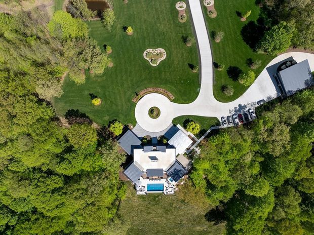 Aerial top-down view of a substantial residential property set within extensive landscaped grounds and mature trees. The image shows a light-coloured multi-wing house with dark pitched roof sections, a blue swimming pool, broad pale concrete driveways, a circular turning court, landscaped garden beds, and a separate outbuilding or garage with parked vehicles nearby.