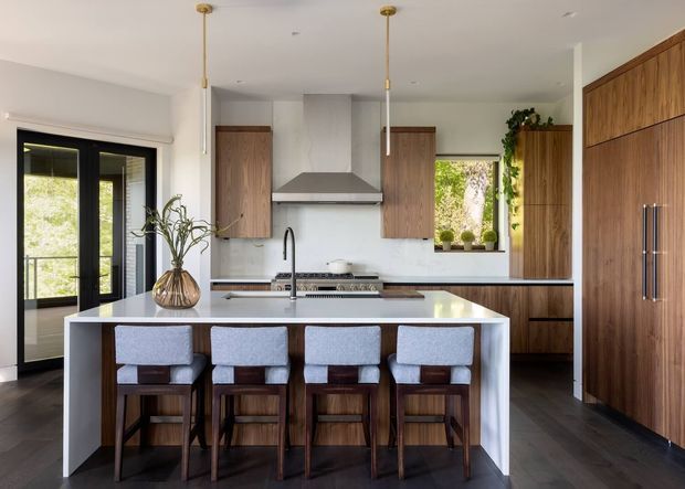 A spacious contemporary kitchen with a large white island and four upholstered timber bar stools. The room features extensive warm timber cabinetry, a dark wood-look floor, white walls and benchtops, black-framed glazed doors, a stainless-steel rangehood, and slim pendant lights. Potted greenery and daylight add a natural, refined feel.