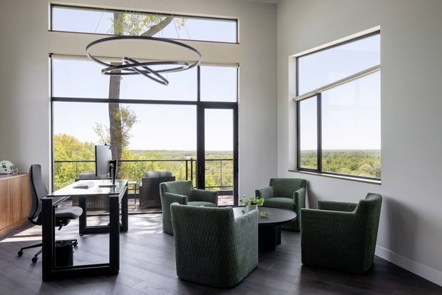 A bright, modern home office or study lounge with floor-to-ceiling black-framed glazing, a desk and office chair, four deep green upholstered lounge chairs around a round coffee table, and a sculptural circular pendant light. The room overlooks a leafy landscape and has dark timber-look flooring with white walls and a high ceiling.
