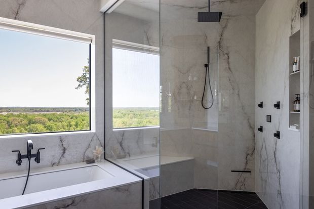 Contemporary bathroom with a white built-in bathtub beside large windows, a frameless glass walk-in shower, matte black shower and tapware, dark floor tiles, and dramatic grey-veined stone-look wall surfaces. The room overlooks a leafy landscape.