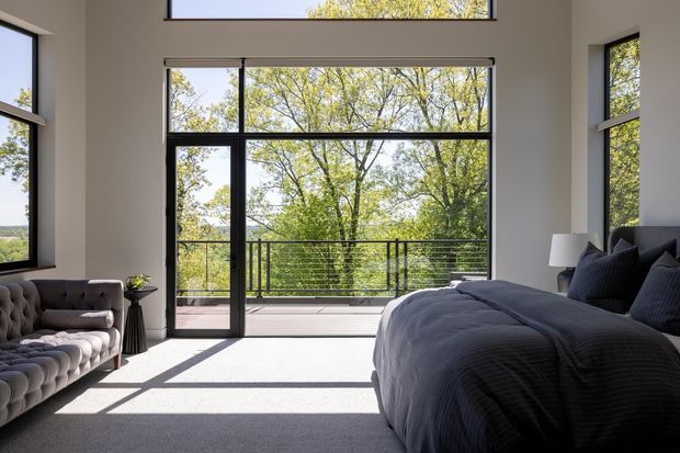 A bright, spacious bedroom with a large upholstered bed, tufted sofa, bedside table and lamp. Floor-to-ceiling black-framed glazing and a glazed door open onto a balcony or terrace overlooking leafy trees, with strong sunlight casting geometric shadows across the carpet. The room has a clean, refined contemporary aesthetic and a high ceiling.