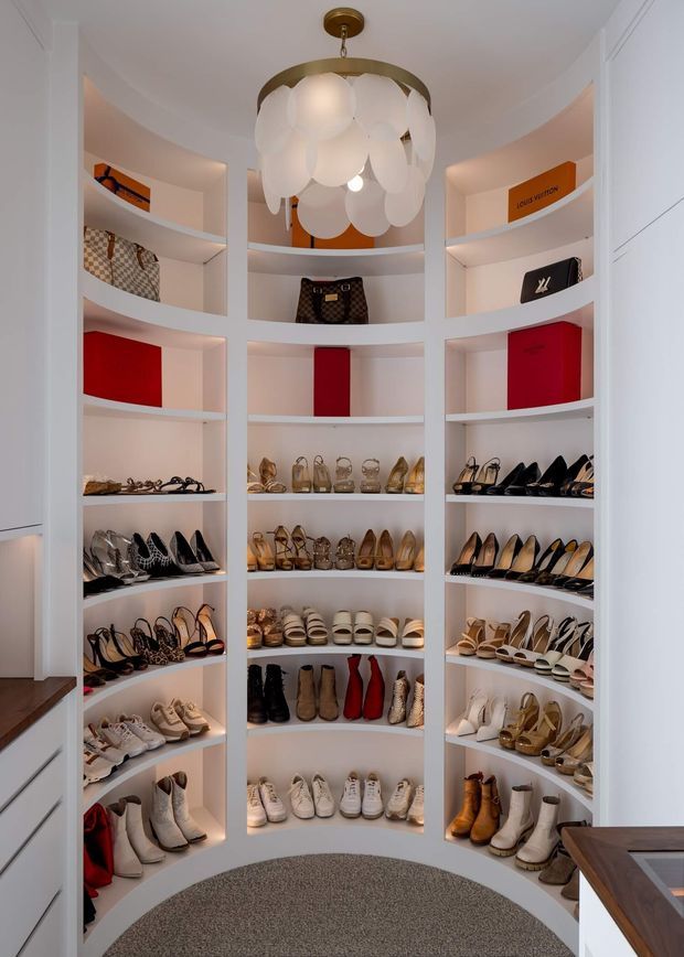 A compact, highly organised walk-in wardrobe with curved white built-in shelving arranged around the room, displaying a large collection of shoes, handbags, and luxury-brand boxes. The space has a softly lit white interior, a decorative pendant light with layered white discs, warm timber countertop edges, and a grey carpeted floor.
