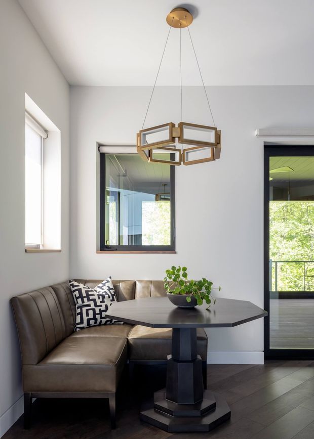 A bright, refined dining nook with a built-in brown leather banquette wrapping around a dark geometric pedestal dining table. A black-and-white patterned cushion and potted plant add contrast. The room has light grey walls, dark timber flooring, black-framed windows and glazed doors, and a suspended geometric brass-and-glass pendant light. Trees and an outdoor balcony are visible through the glazing.
