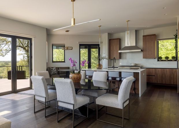 A bright, open-plan kitchen and dining area with a large island, dark timber cabinetry, integrated stainless-steel appliances, and a formal dining table with upholstered chairs. Full-height glazed doors open to a balcony or terrace, while large windows provide views of greenery. The room has recessed ceilings, slim contemporary pendant lighting, dark timber flooring, and a neutral grey-beige palette.
