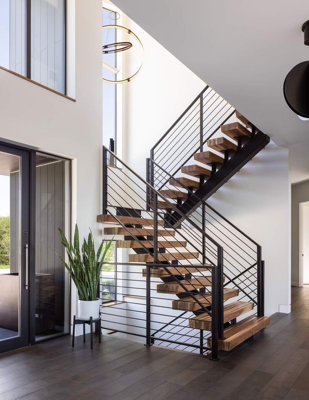 Bright contemporary residential entryway dominated by an open-riser staircase. The staircase has dark metal stringers and slim horizontal cable railings with warm timber treads, set against white walls and tall glazing. A dark timber-look floor, glass entry door, potted plant, and sculptural pendant light complete the clean, airy space. A hallway and upper-level void are visible beyond the stair.