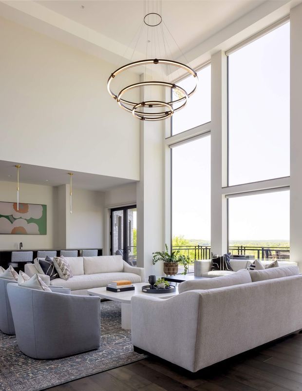 Bright, spacious contemporary living room with a double-height ceiling and floor-to-ceiling windows overlooking a distant landscape. The room features large pale upholstered sofas and curved armchairs arranged around a low coffee table, a patterned area rug, dark timber flooring, minimalist artwork, indoor plants, and a striking multi-tiered circular pendant light. An open kitchen or dining area is partially visible in the background.
