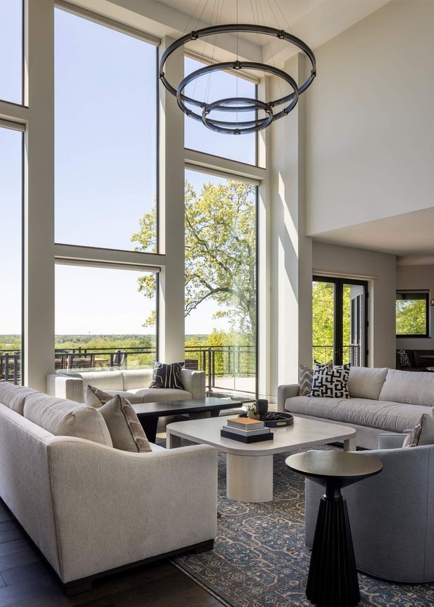 A refined contemporary living room with multiple pale upholstered sofas and armchairs arranged around sculptural coffee tables. The room features dramatic floor-to-ceiling windows, a high ceiling, expansive views, dark timber flooring, a patterned area rug, and a large multi-ring pendant light. Neutral furnishings and white architectural surfaces create a bright, spacious atmosphere.