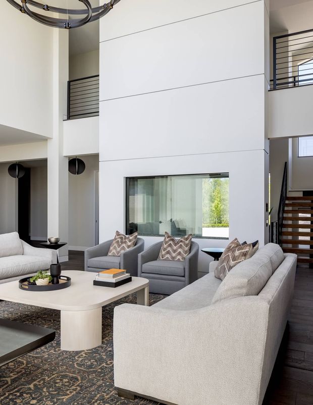 A spacious double-height living room with a neutral upholstered seating arrangement, including a large curved sofa and matching armchairs around a light coffee table. The room features a patterned area rug, contemporary circular chandelier, white paneled walls, internal horizontal metal railings, a large glazed opening, and a staircase leading to an upper level.