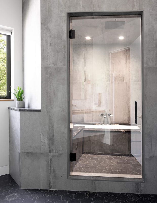 A contemporary bathroom featuring a fully enclosed glass shower with black metal framing and hardware. Large grey concrete-look wall tiles surround the shower and vanity area, while dark hexagonal floor tiles extend across the room. A compact dark stone vanity ledge sits beneath a window, with a small potted plant adding greenery. The shower includes recessed lighting, a ceiling rain shower fixture, wall controls, and a built-in bench or ledge.