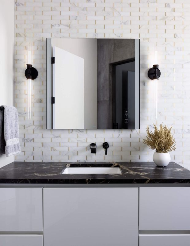 Contemporary bathroom vanity featuring a large frameless mirrored cabinet, white vertically and horizontally patterned wall tile with subtle gold accents, a dark marble-look benchtop, white integrated basin, matte white vanity cabinetry, and black wall-mounted tapware and sconces. A towel and small decorative vase are visible at the edges.