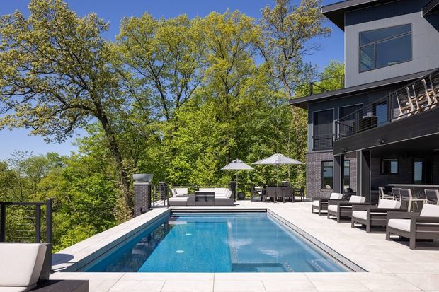 A modern multi-level home with dark cladding, extensive glazing, and a large elevated outdoor terrace surrounding a rectangular swimming pool. Contemporary outdoor lounge and dining furniture, umbrellas, and a glass or dark-framed balustrade overlook a leafy wooded setting. The scene is sunny, refined, and resort-like.