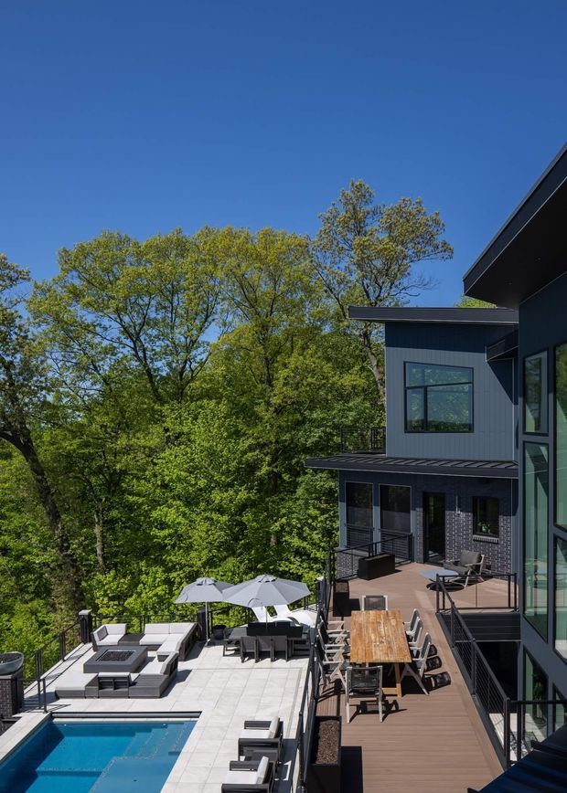 A contemporary multi-level dark-clad home set among mature trees, viewed from an elevated exterior position. The image shows extensive outdoor entertaining areas including a timber deck with a long dining table, a paved terrace with outdoor lounge furniture and umbrellas, and a partially visible swimming pool. Large black-framed windows and projecting flat roof forms define the building facade.