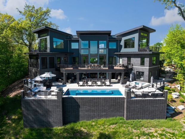A large multi-storey contemporary dark-clad residence viewed from the rear, set among mature trees. The home has several projecting upper-level volumes with low-pitched roofs, extensive glazing, broad terraces and balconies, and a landscaped outdoor entertaining area with a swimming pool, outdoor seating, umbrellas, and dining furniture.