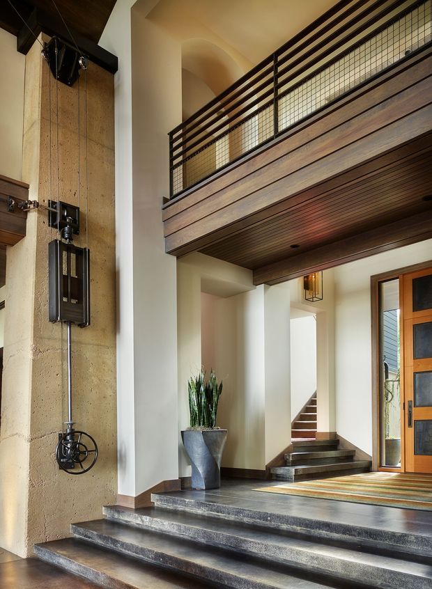 A grand residential entry foyer with broad dark stone steps leading to a timber-framed front door. The double-height space features a mezzanine balcony with dark timber cladding and black metal-and-wire balustrades, a tall textured stone or concrete feature wall with exposed mechanical hardware, white plaster columns, timber-lined soffits, recessed stair access, and a sculptural planter. Warm natural materials and strong vertical and horizontal lines create a refined, dramatic arrival space.