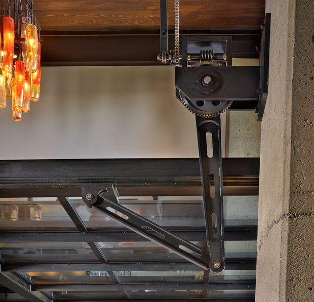 Close-up architectural detail of an exposed industrial-style overhead mechanism and structural framing. A large black metal pivoting arm and toothed gear assembly is mounted against a textured concrete wall, with dark steel beams and glazed panels below. Warm wood ceiling cladding and amber glass pendant lights add contrast to the utilitarian structure.