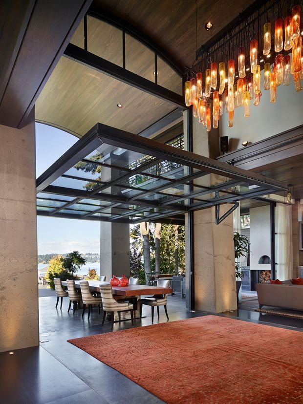 A dramatic open-plan interior combining a dining area and living room beneath a high, partially double-height ceiling. Large concrete columns, dark timber wall and ceiling finishes, black steel framing, and extensive glass overhead create a strong indoor-outdoor connection with views of a waterfront landscape. A large sculptural cluster pendant light hangs above the living area, while a long dining table and upholstered chairs sit near the glazing. A warm red-orange area rug adds contrast to the dark floor.