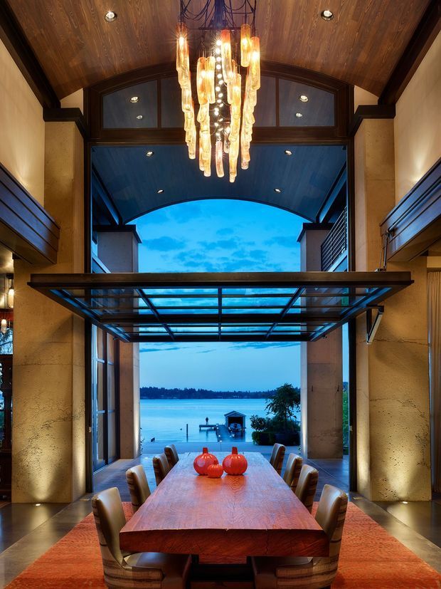 A dramatic dining room centered on a long solid timber dining table with upholstered dining chairs and a large sculptural glass pendant chandelier. The room features dark timber ceiling panelling and trim, warm textured wall surfaces, extensive glazing, and a retractable or glazed roof opening leading toward a waterfront view and private jetty. The composition emphasizes a strong indoor–outdoor connection and evening blue-hour atmosphere.