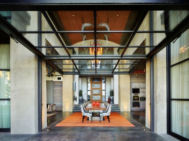 View through a dramatic glazed entry or breezeway into a refined dining area. The space features exposed concrete walls, dark metal-framed floor-to-ceiling glazing, a transparent glass ceiling, warm timber ceiling panels, polished dark flooring, and a long dining table centred on an orange rug. A contemporary kitchen or service joinery area is visible beyond the dining room.
