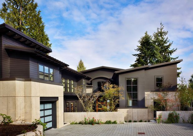 Exterior view of a contemporary single-storey residential home composed of several connected wings. The facade combines dark horizontal cladding, pale concrete or stone-like lower walls, large rectangular windows, and a low-pitched roof with broad eaves. A paved courtyard or driveway occupies the foreground, with mature trees and planted garden beds around the building.