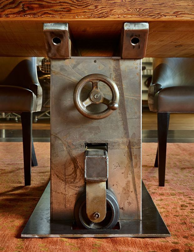 Close-up interior detail of a custom industrial-style metal support or console beneath a substantial timber tabletop. The distressed steel panel features a prominent vintage handwheel, latch, and caster-like hardware, with leather-upholstered dining or lounge chairs positioned on either side over a patterned carpet.
