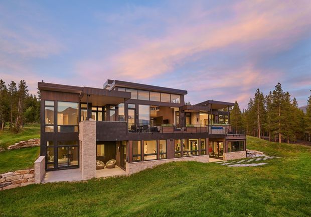 A large contemporary multi-level house set in a forested landscape, photographed from the exterior at dusk. The home features extensive floor-to-ceiling glazing, dark metal-framed windows, broad cantilevered roof planes, stone-clad structural piers, multiple terraces and balconies, and a mix of horizontal and vertical forms. A broad lawn and surrounding coniferous woodland frame the building.