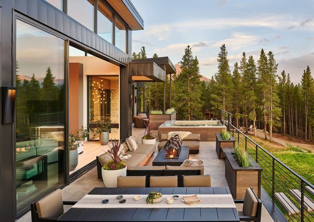A contemporary multi-level home with a spacious timber-toned terrace overlooking a forested landscape. The outdoor living area includes a dining table, lounge seating, built-in planters, a linear fire feature, and a raised spa or plunge pool. Large floor-to-ceiling glazed openings connect the terrace to the interior, while dark metal railings and crisp horizontal cladding define the modern facade.