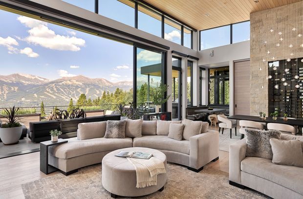 A spacious, high-end open-plan living room with panoramic floor-to-ceiling glazing overlooking a dramatic mountain landscape. The room features a large curved neutral sectional sofa, matching armchair, round upholstered ottoman, dark side tables, a light stone or textured feature wall with integrated wine storage, and a warm timber-lined ceiling. The interior is bright, refined, and contemporary, with strong indoor-outdoor visual connection.