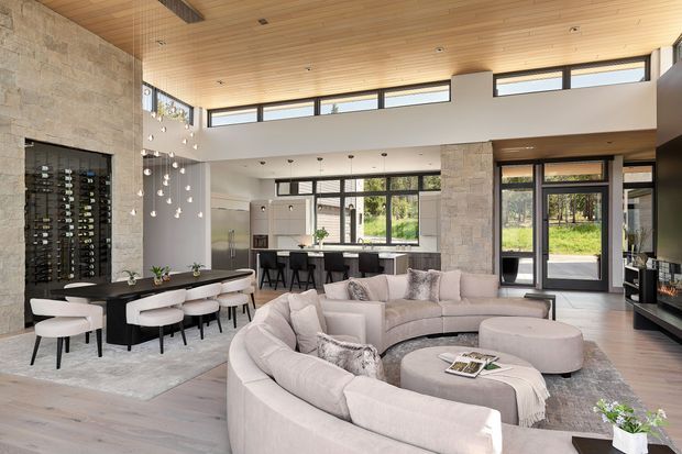 Spacious open-plan living, dining and kitchen area with a double-height ceiling and clerestory glazing. The room features a large curved light-coloured sectional sofa, black dining table and chairs, a stone-clad feature wall with integrated wine display, a pale stone kitchen island, timber ceiling lining, and extensive dark-framed windows and doors. A mezzanine-like upper level and suspended decorative pendant lights add visual interest.