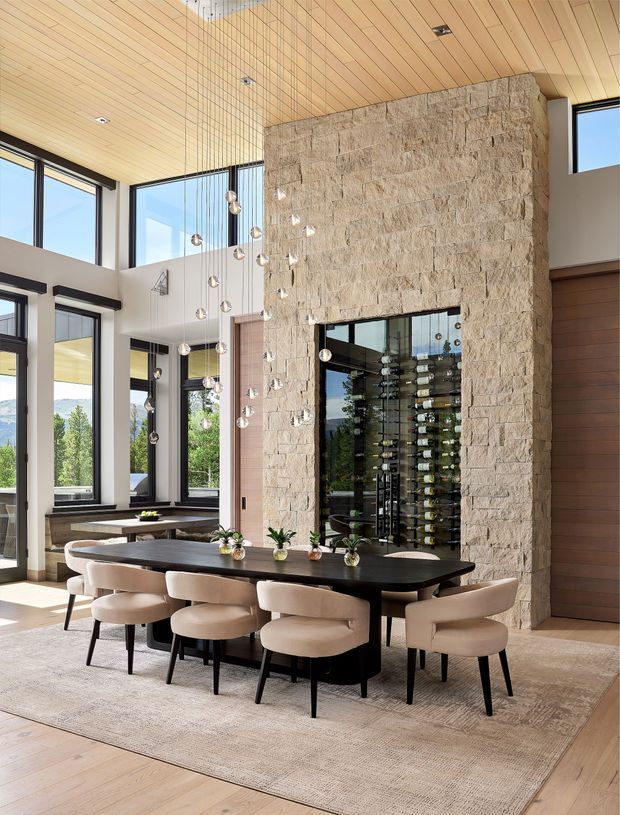 Bright, double-height dining area with a long dark dining table and upholstered chairs, framed by expansive black-framed glazing. A dramatic rough-stone feature wall contains a glass-fronted wine display, while a timber-lined ceiling, suspended pendant cluster, and warm timber finishes create a refined contemporary interior.