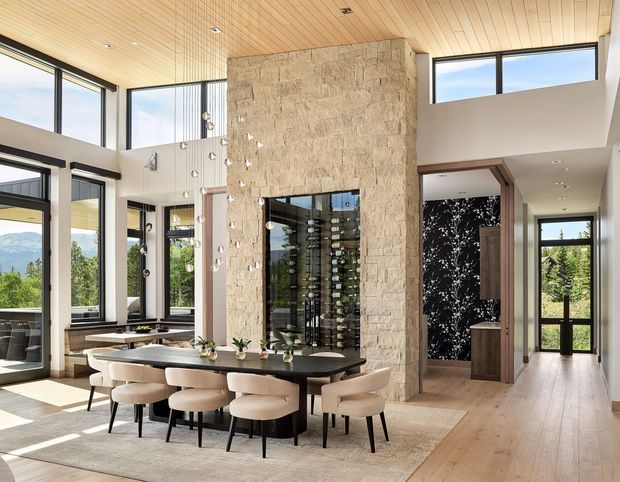 A bright, spacious open-plan dining and living area with a long dark dining table surrounded by upholstered chairs. A dramatic full-height natural stone feature wall incorporates a glass-fronted wine display, while expansive black-framed glazing, a timber-lined ceiling, pale timber flooring, and a tall entry hall create an airy contemporary interior. Mountain and garden views are visible through the windows.