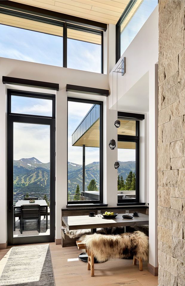 A bright double-height dining area with floor-to-ceiling black-framed glazing overlooking a dramatic mountain landscape. The space features a light timber ceiling, pale timber flooring, white walls, exposed stone masonry, a dark timber dining table, and rustic sheepskin-covered stools. Pendant lights hang in the tall void, and a small dining setting is visible through the glazed door.