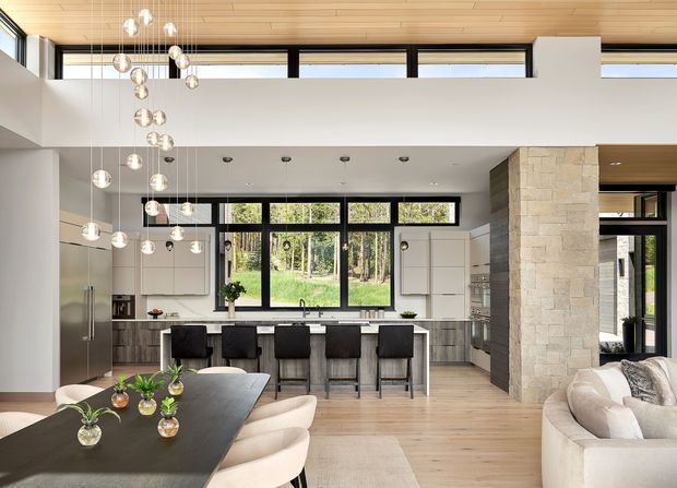 A bright, open-plan contemporary kitchen, dining, and living area with a double-height ceiling. The kitchen features a central island with multiple bar stools, white flat-panel cabinetry, dark benchtops, and large black-framed windows overlooking trees. A substantial pale stone-clad structural column separates the kitchen from the living zone. The dining area has a long dark table with curved upholstered chairs, while the living area contains a light sectional sofa. Numerous suspended glass pendant lights create a prominent vertical feature, complemented by timber ceiling lining and light timber flooring.