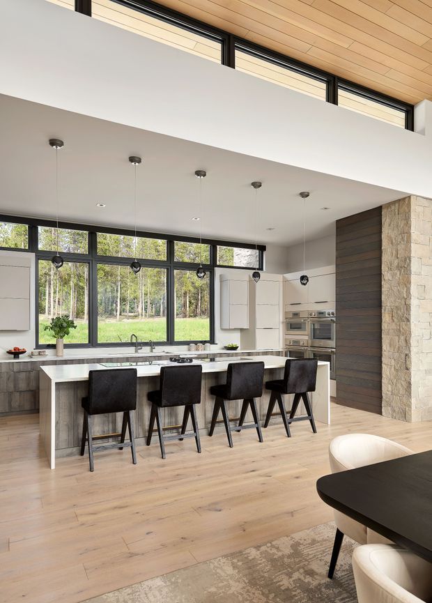 A bright, spacious contemporary kitchen with a large white island seating four, dark upholstered bar stools, extensive black-framed glazing, white cabinetry, integrated stainless-steel appliances, and a timber-lined ceiling. A stone-clad column and dark horizontal timber wall create contrasting feature surfaces. The kitchen opens toward a dining area in the foreground, with a forested landscape visible through the windows.