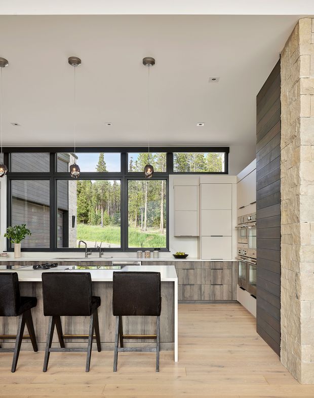 A contemporary kitchen with a large island and two dark upholstered bar stools. The room features light timber flooring, wood-grain cabinetry, white integrated cabinetry, dark-framed floor-to-ceiling windows, pendant lighting, and a prominent natural stone-clad structural wall. Views of trees are visible through the glazing.