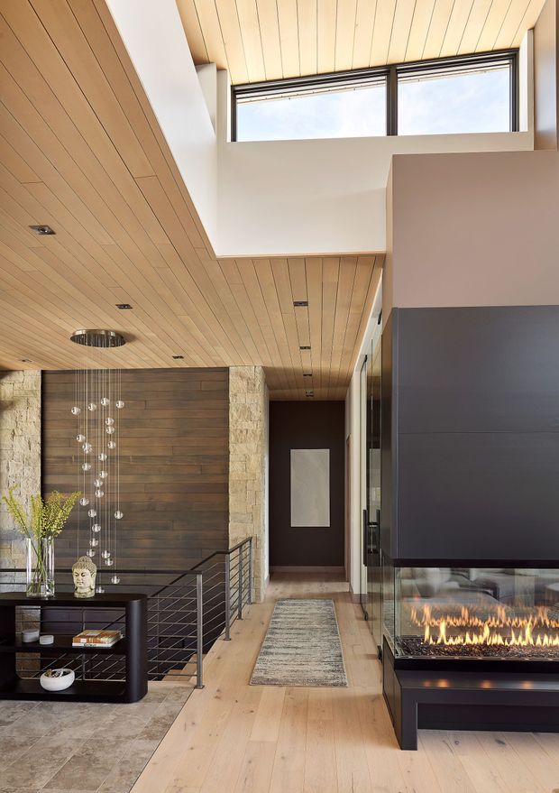 A sophisticated open-plan residential interior viewed from a hallway or entry area, featuring a double-height void with clerestory glazing, extensive warm timber plank ceilings, pale timber flooring, a dark timber feature wall, exposed light stone, black metal balustrading, and a contemporary built-in fireplace with visible flames. A sculptural pendant hangs over the stairwell, while a console and decorative objects occupy the foreground.