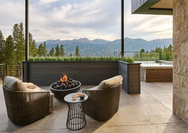 A contemporary outdoor terrace overlooking a dramatic mountain range. The covered patio features two woven lounge chairs arranged around a round fire bowl and small side table, with a raised dark planter, glass and metal balustrade, and an integrated outdoor kitchen or counter area. Stone-clad walls and a timber-lined roof overhang frame the view.