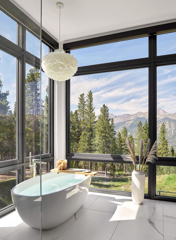 Bright, contemporary bathroom with a freestanding white bathtub positioned beside expansive black-framed floor-to-ceiling windows. The room has large light marble-look floor tiles, white walls, a sculptural pendant light, and a tall white vase with dried grasses. Mountain and forest scenery is visible beyond the glazing.