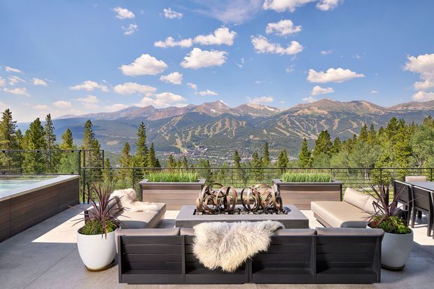 A contemporary rooftop or elevated outdoor terrace with panoramic mountain views. The space features a large modular outdoor lounge arrangement around a central rectangular fire table, with neutral upholstered seating, dark horizontal-panel cladding, sculptural metal decor, potted ornamental plants, and a glass-and-metal balustrade. Bright sunny conditions and the dramatic alpine landscape create an inviting resort-like setting.