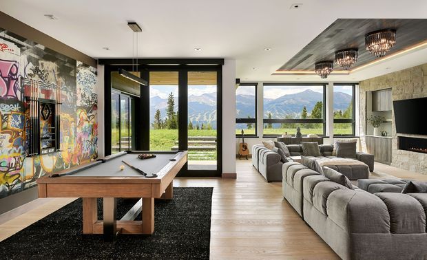 A spacious contemporary living and recreation room with floor-to-ceiling glazing overlooking a mountainous landscape. The room features a large grey sectional sofa, pool table, timber flooring, a graffiti-style feature wall, stone-clad fireplace wall, and a dramatic recessed ceiling with multiple pendant lights.