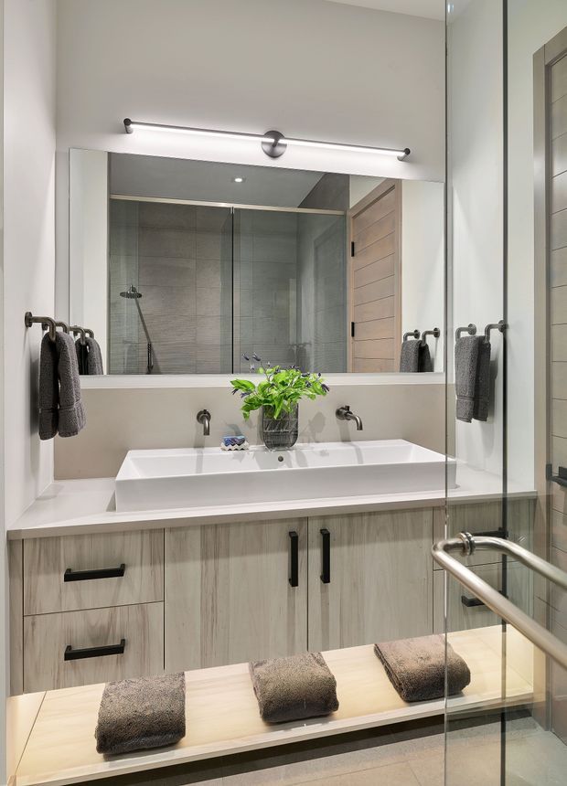 A contemporary bathroom focused on a wide double vanity with a white integrated basin, pale timber-look cabinetry, black hardware, and a large wall mirror. A glass-enclosed shower with grey tiled walls is visible behind the vanity, along with towel rails, a linear vanity light, and a timber door. The room has a clean, refined, neutral finish with warm wood accents.