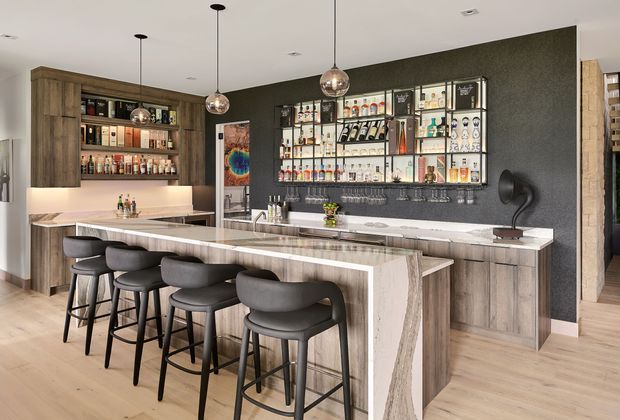 A sophisticated residential home bar with a large stone-topped island and seating for several people. Timber cabinetry and open shelving display bottles and glassware, while a dark textured feature wall supports illuminated display shelving. Three pendant lights hang above the island, complemented by pale timber flooring, integrated appliances, and a compact sink area.