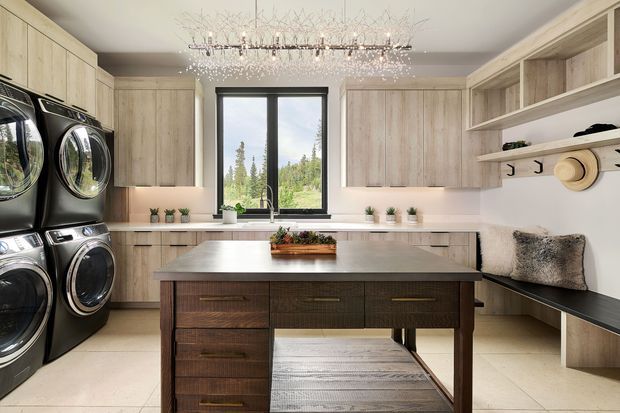 A bright, spacious kitchen-laundry room with pale timber cabinetry, a large central island, integrated bench seating, open shelving, and a stacked bank of front-loading appliances. A large window brings in natural light and views of trees. The room combines warm wood tones, white walls, light flooring, dark stone-like benchtops, and a decorative chandelier.
