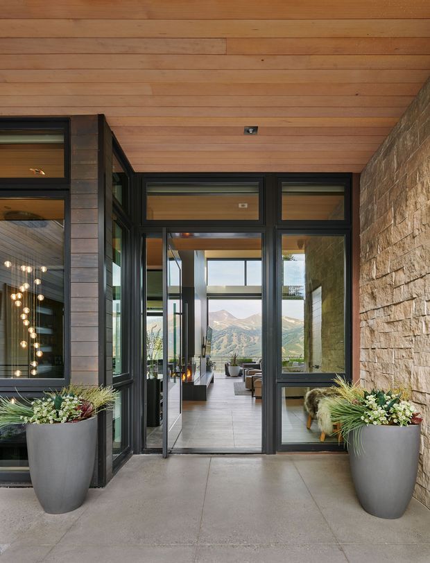 A covered residential entry or transition space with warm timber soffit lining, dark framed glazing and doors, a textured natural stone wall, and large planters. The open glazed doorway leads into a bright interior living area, with a mountainous landscape visible beyond.