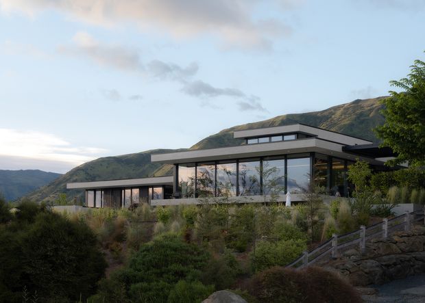 A low-profile contemporary hillside residence with broad flat roof planes, extensive floor-to-ceiling glazing, and dark horizontal framing. The house is integrated into a landscaped, sloping site with native-style planting, stone retaining walls, and mountain terrain behind it.