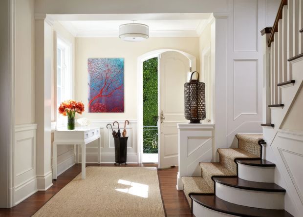 Bright, refined residential entryway with cream-painted panelled walls, white trim, a narrow console table, a patterned artwork, an open front door, and a curved staircase with carpeted treads and dark timber nosings. Warm timber flooring and a woven runner create a welcoming transitional interior.