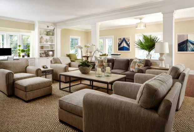 A spacious, traditionally detailed living room with multiple upholstered sofas and armchairs arranged around a large rectangular coffee table. The room features warm beige walls, white architectural trim and columns, built-in open shelving, layered neutral furnishings, framed artwork, indoor plants, and patterned carpeting. Large windows provide abundant natural light, creating a refined and inviting atmosphere.