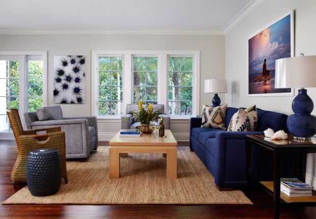 Bright, traditionally detailed living room with pale walls, white-trimmed multi-pane windows, a navy sofa, grey armchairs, wicker seating, a light timber coffee table, and a large natural woven rug. Blue ceramic lamps and coastal-themed artwork add colour, while leafy greenery is visible through the windows.