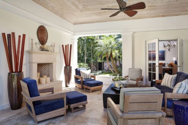 Bright, spacious living room with a traditional fireplace, pale wood-look ceiling, light neutral walls, stone-tile flooring, and multiple light timber-framed lounge chairs upholstered in deep blue and neutral fabrics. A large opening overlooks lush tropical planting, while French doors lead to an adjacent room. Decorative vases, a coffee table, and a ceiling fan complete the relaxed, refined interior.