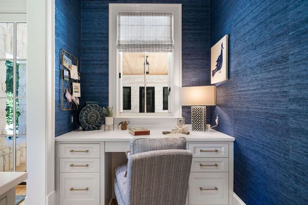 Compact, refined home office nook with a built-in white desk and drawer cabinetry set against richly textured deep blue walls. A central window with a white frame and blind provides natural light, while a padded desk chair, table lamp, artwork, and small desk accessories complete the space. A glazed opening at left reveals greenery and an adjacent bright room.